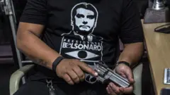 Um cliente vestindo uma camisa em apoio a Jair Bolsonaro manuseia uma arma de fogo à venda em uma loja de armas em São João do Meriti, em 14 de janeiro de 2018.