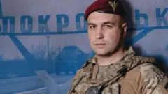 Євген Ласійчук у формі десантника, на фоні стели з написом Покровськ