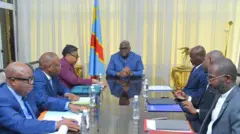 Perezida wa DR Congo Félix Tshisekedi yicaye mu nama akikijwe ku meza n'abategetsi bakuru barimo na Minisitiri w'intebe Judith Suminwa. Inyuma ya Tshisekedi hari igitambaro cyo gukingira idirishya cy'ibara rya khaki rijya kuba umuhondo hamwe n'ibendera rya DR Congo ry'ubururu, umuhondo n'umutuku. Ifoto yatangajwe n'ibiro bya Perezida Tshisekedi, ku wa kane, ku itariki ya 11 Ukuboza (11) mu mwaka wa 2025. 