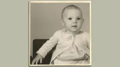 Uma foto em preto e branco do pai de Matthew quando era bebê, na década de 1940. Ele está usando um cardigã branco de tricô e olhando para fora da câmera, à direita