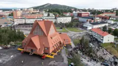 crkva u švedskom gradiću iz vazduha, oko nje se vidi velika grupa ljudi kako je okružuje, a u pozadini su duge zgrade, kuće i planina.
