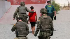 Três soldados e um agente da Guarda Civil acompanham um menor vestindo uma camiseta vermelha e bermuda. Todos estão de costas. Ceuta, 1º de agosto de 2026.