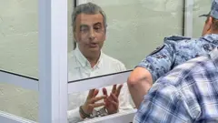 手錠をかけられた白髪の男性が、法廷のガラス張りのおりの中に座り、話している