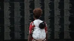 Umugore uhetse umwana ari ku rwibutso areba amazina y'abazize jenoside yabaye mu Rwanda mu 1994