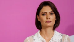 Michelle Bolsonaro olhando para cima durante evento, com olhar sério, e painel rosa atrás