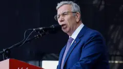 Lacivert takım elbiseli, beyaz saçlı bir adam mikrofonda konuşuyor