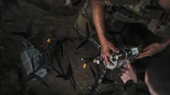Un brazo sostiene un dron de color plateado posado en el suelo en la oscuridad.