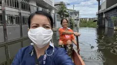 Seorang perempuan yang mengenakan masker, menumpang sebuah sampan untuk mengarungi jalanan yang tergenang oleh banjir.
