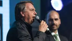 Sorrindo, Eduardo Bolsonaro observa seu pai discursando