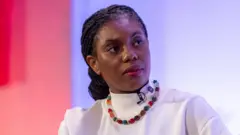 Kemi Badenoch wear one white shirt and multicoloured necklace. Dem clip microphone to her neckline and she stand behind one red and purple background.