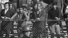 Fotografía en blanco y negro en la que se ve a personas detenidas en un estadio deportivo tras el golpe de Estado. Un soldado armado con un rifle se encuentra de pie frente a la valla, mientras los prisioneros esperan a ser interrogados.