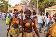 Deux jeunes filles dans de tenues traditionnelles, elles tiennent de longs bâtons à la main droite et sourient dans la caméra. D'autres jeunes gens en file indienne sont en arrière-plan.