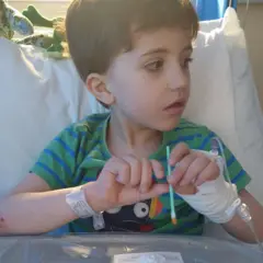 Criança sentada na cama, com uma mão enfaixada e um tubo médico, segurando um pequeno swab