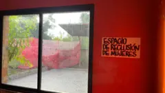 Otra imagen del interior del memorial. En ella se ve una gran ventana con un cartel junto a ella, colgado en una pared de color rojo vivo, en el que se lee "Espacio de reclusión de mujeres".