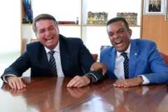 De terno preto e gravata listrada, Bolsonaro, está sentado ao lado de Otoni de Paula, que usa terno azul, gravata estampada e segura o braço do então presidente. Ambos gargalham.