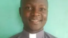 Padiri Deusdedit Ssekabira yambaye ishati isa n'ivu iriho n'ikora ry'umweru riranga abapadiri. 