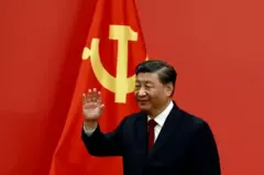 Ifoto ya Prezida w'Ubushinwa Xi Jinping, azamuye ikiganza. Yambaye ikote ry'umukara.