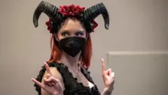 One woman wear one black horn and covid face mask and give sign of di horn