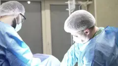 На фото крупним планом двоє хірургів у синьому спецодязі схилилися під час проведення операції військовому. 