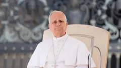 O Papa Leão 14 faz seu discurso aos fiéis durante a Audiência Geral de quarta-feira, na Praça de São Pedro, no Vaticano, em 5 de novembro de 2025. Ele veste sua tradicional túnica branca e está sentado em uma cadeira de cor creme. Atrás dele, vê-se um fundo desfocado da fachada ornamentada da Praça de São Pedro.