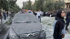 Una mujer con pañuelo en la cabeza se encuentra a la derecha de un auto dañado y cubierto de polvo.