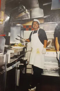 Hombre con delantal trabajando en una cocina industrial, usando pinzas frente a una parrilla con comida, rodeado de equipos de metal.