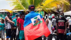 Um migrante haitiano com a bandeira nacional do Haiti, enquanto milhares de migrantes haitianos, africanos e cubanos tentam atravessar para a fronteira entre a Colômbia e o Panamá de barco, com o objetivo de seguir rumo aos Estados Unidos.