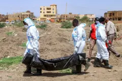 Dos hombres con ropas de protección blanca cargan una bolsa negra con un cadáver en medio de un terreno excavado. Hay otros tres hombres en la escena.