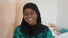 Fatou Tall Mbow, porte un voile noir et un tissu vert et noir. Elle sourit face caméra.