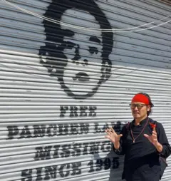 Homem usando roupas pretas e uma faixa vermelha em frente a uma imagem de Panchen Lama pintada na porta de uma loja
