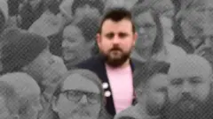 Uma imagem dessaturada de uma multidão, com Stephen McCullagh no centro destacado em cores. Ele veste uma jaqueta escura e uma camiseta rosa