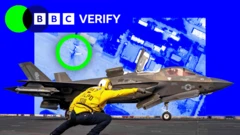 A US navy airman gives an F-35 fighter a signal to take off. He is wearing a yellow jacket and a helmet and is pointing towards the runway. The jet is armed with missiles and is painted grey. The image is imposed over a satellite photo of an airbase in Iran where damaged jets are highlighted. 