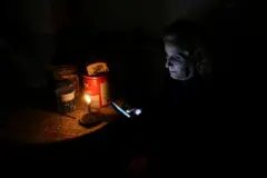 Uma mulher está sentada à mesa, o rosto iluminado por uma vela, segurando um celular nas mãos.