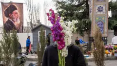 A flower vendor holds purple and white flowers over his face as people shop in preparation for Nowruz celebrations on 19 March 2026 in Tehran, Iran. Behind him on the left, a poster of former Supreme Leader Ayatollah Ali Khamenei, who was killed in the first wave of US-Israeli strikes, is displayed. A few people walk behind the vendor. 