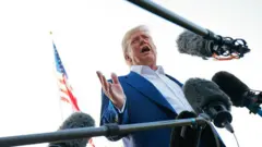 Imagem de Donald Trump vestido com paletó e camisa de colarinho aberto, cercado por microfones. A imagem foi tirada de baixo para cima.