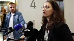 Elisabeth Guthmann, vestida con un abrigo oscuro y gafas, habla por los micrófonos.