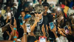 Umuhanzi w’umugabo yambaye amarori n’inkofero arimo kuvugira muri ‘micro’. Akikujwe n’abantu benshi, bamwe bafite telefone ngendanwa bariko baramufotora. 