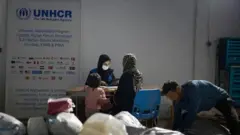 یک کارمند زن ملل متحد در تصویر واضح است که پشت ی میز نشسته و با یک خانم و دخترش صحبت می‌کند. یک مرد هم در گوشه راست تصویر دیده می‌شود.