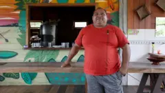 Roberto Brito, um homem corpulento vestindo camiseta vermelha, posacomo funciona o cbetfrente a uma mesacomo funciona o cbetmadeira e a uma construção tambémcomo funciona o cbetmadeira, pintada com um desenho coloridocomo funciona o cbetfolhas amazônicas