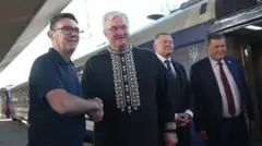 Burnham ściska dłoń Sybihy, gdy oboje się uśmiechają i patrzą przed siebie, stojąc obok dwóch innych mężczyzn i pociągu