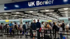 Aworan yii ṣafihan awọn alawọ funfun lẹnu ibode abawọle UK. Pupọ wọn pọn baagi sẹyin, wọn si fi ibomu bo imu wọn. Ina tan lati oke abawọle naa, bẹẹ ni wọn fi awọn okun dudu dá aaye ti awọn arinrinajo to fẹẹ wọle naa duro si. Akọle to kede ẹnu ibode naa han loke pẹlu.