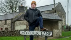 A smiling man in a raincoat and yellow hat stand with one foot resting on a road sign that says "Mycock Lane"