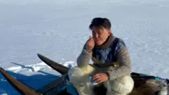 Un hombre sentado sobre un trineo en una superficie helada come con una cuchara.