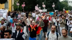 Un numeroso grupo de personas, algunas gritando, otras con los brazos en alto y coreando, participa en una manifestación. Algunas sostienen siluetas de flamencos rosas, el símbolo de la protesta.