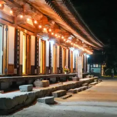 Un templo coreano tradicional iluminado por cálidas luces por la noche, con un tejado curvo de tejas, columnas de madera y escalones de piedra que bordean un amplio patio. Varios visitantes caminan por el sendero que discurre junto al templo.