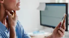 Foto de arquivo de uma mulher gesticulando para o pescoço enquanto segura um telefone em uma videochamada. Ela segura uma caneta na mesma mão. Está sentada em uma mesa, com a tela de um computador atrás dela.