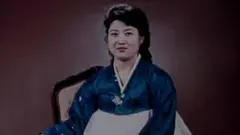 Ko Yong-hui, provavelmente na casa dos 30 ou 40 anos, usa um vestido coreano azul e branco e sorri para a câmera.