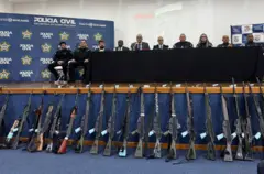 Una serie de rifles capturados por la policía son exhibidos en una conferencia de prensa de la policía civil, mientras los funcionarios hablan detrás.