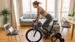 Mujer haciendo ejercicio en una bicicleta fija en la sala de la casa, vistiendo ropa deportiva