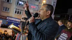 Özgür Özel elinde mikrofonla yüksek bir yerden kalabalığa sesleniyor, geri planda 20 askerin fotoğrafının yer aldığı bir pano tutan bir erkek var. 